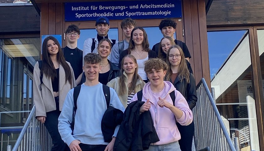 Der Sportkurs aus der KS1 steht vor den Stufen zum Eingang des sportmedizinischen Instituts.